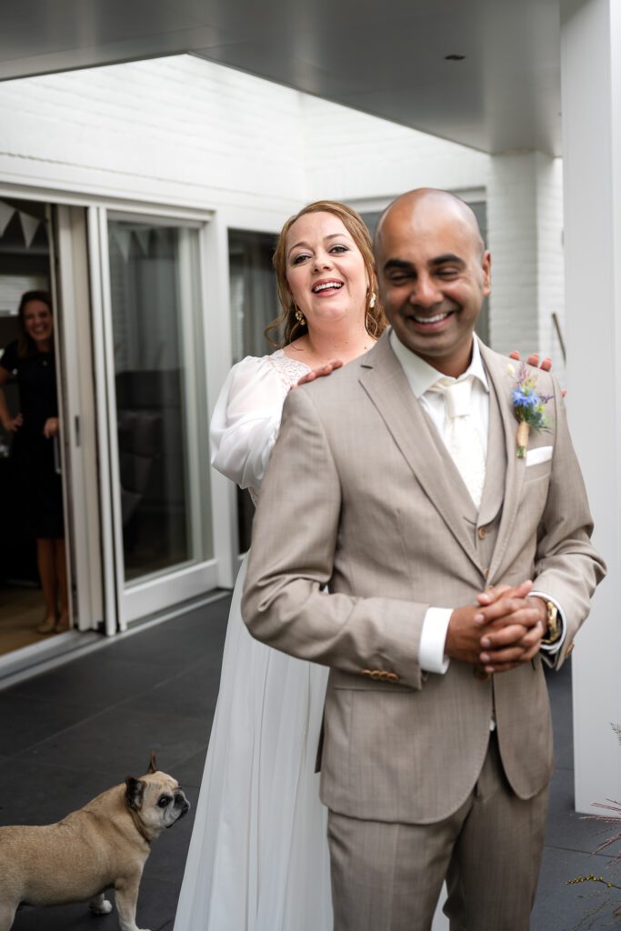 A groom stands smiling with his back to a bride as she taps him on the shoulder for a First Look