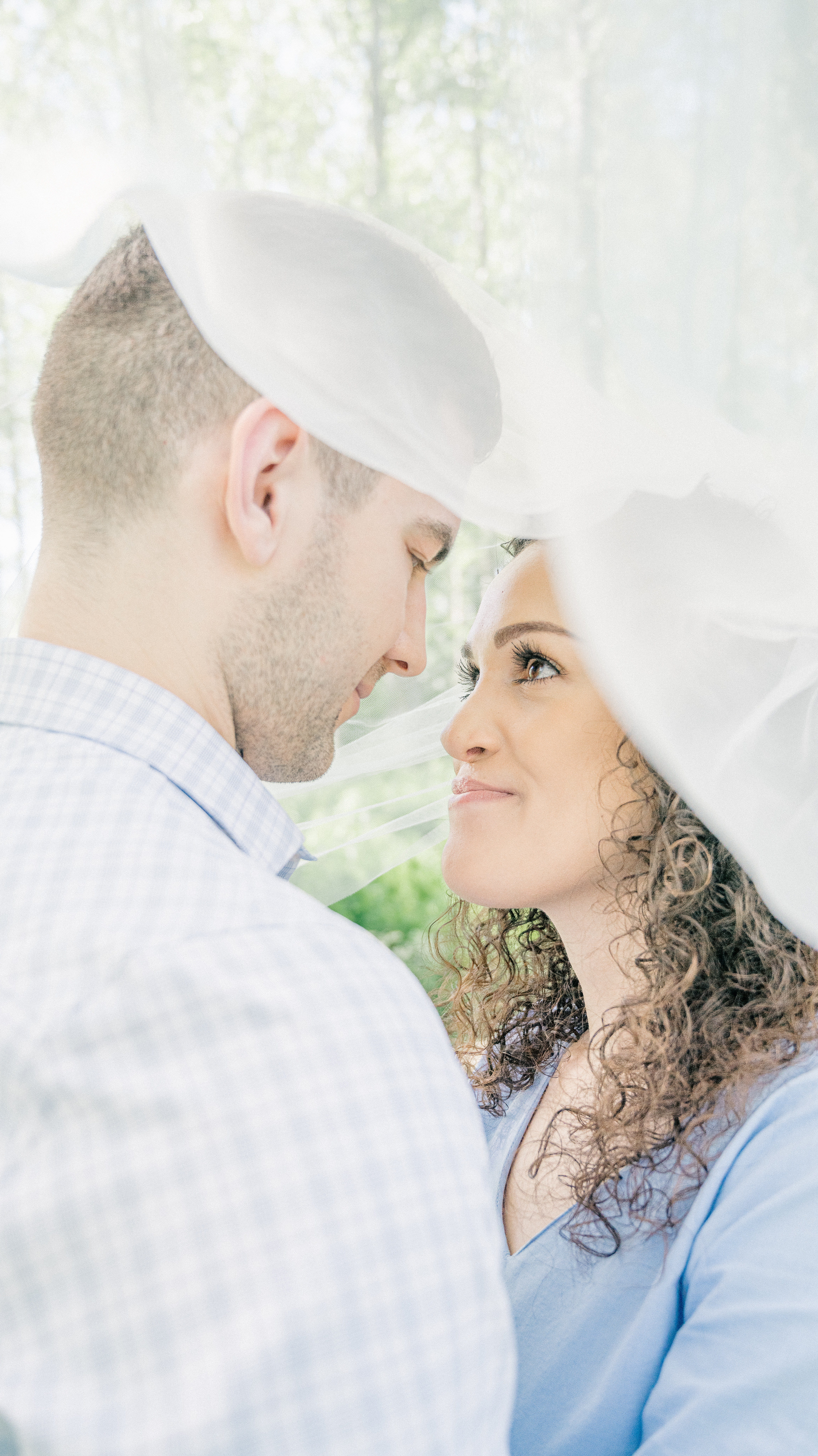 Engagement photo with fiancés looking into each other's eyes under a veil in a lightly wooded green forest