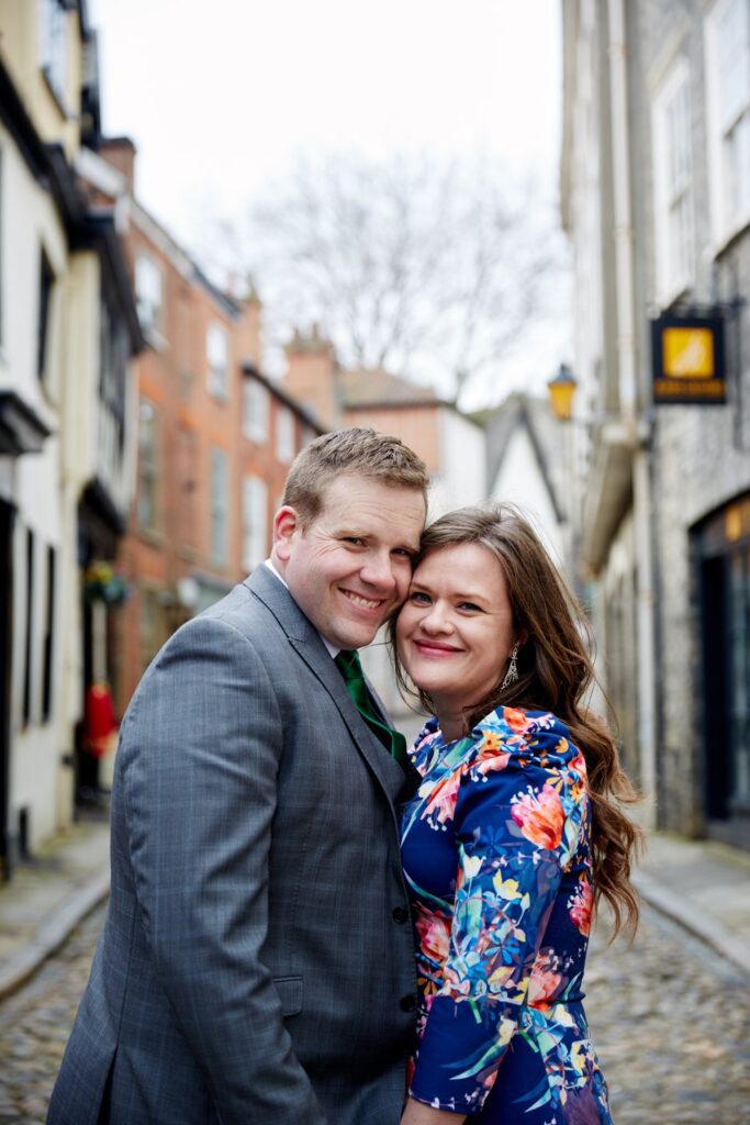 A man and woman stand in a cobbled street Elm Hill in Norwich facing each other with their heads side by side facing the camera