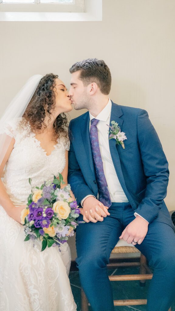 A bride and groom sit and kiss under the original windows of an old church