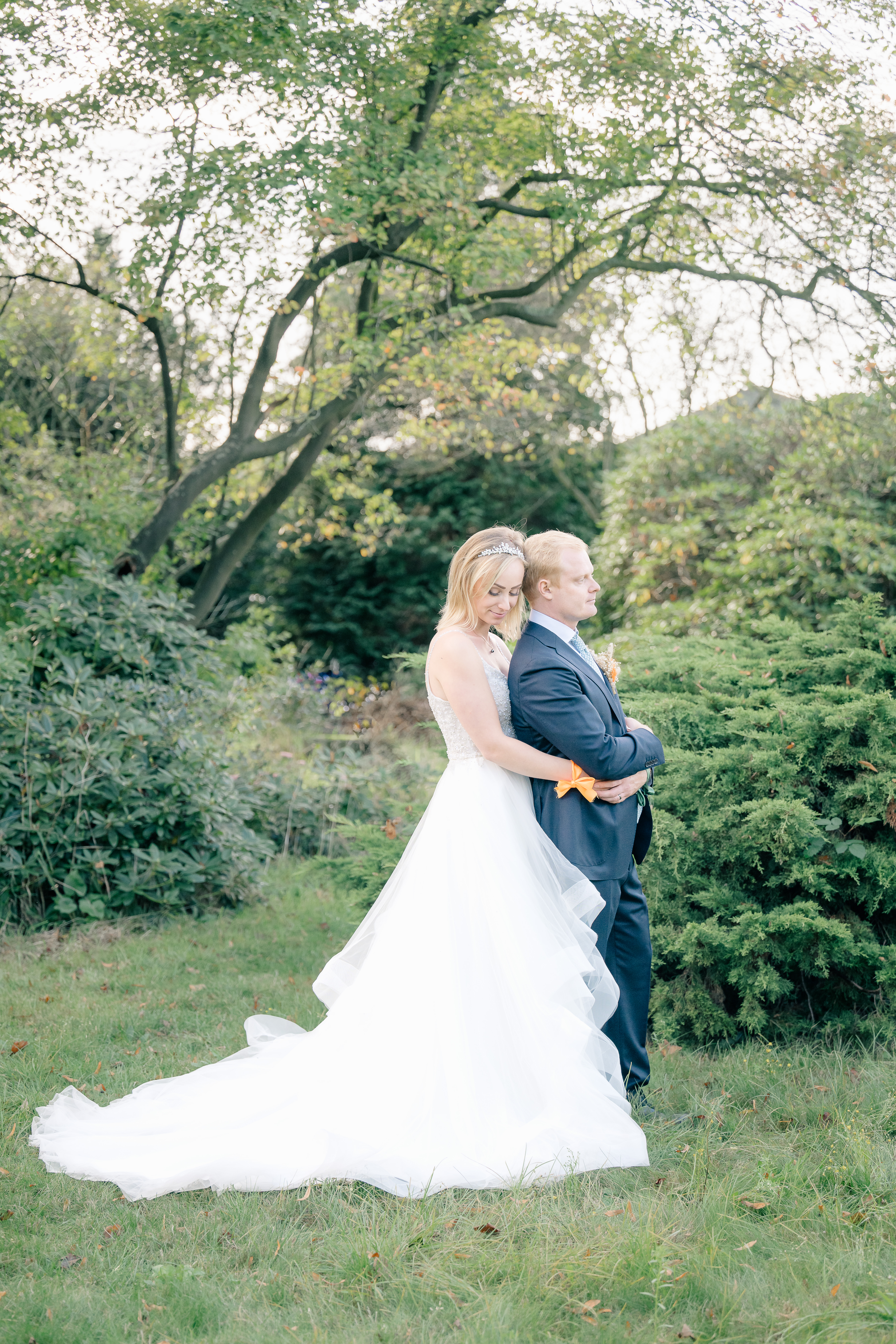 Bride and groom portrait with bride standing behind groom and embracing him while he looks ahead in a garden wedding
