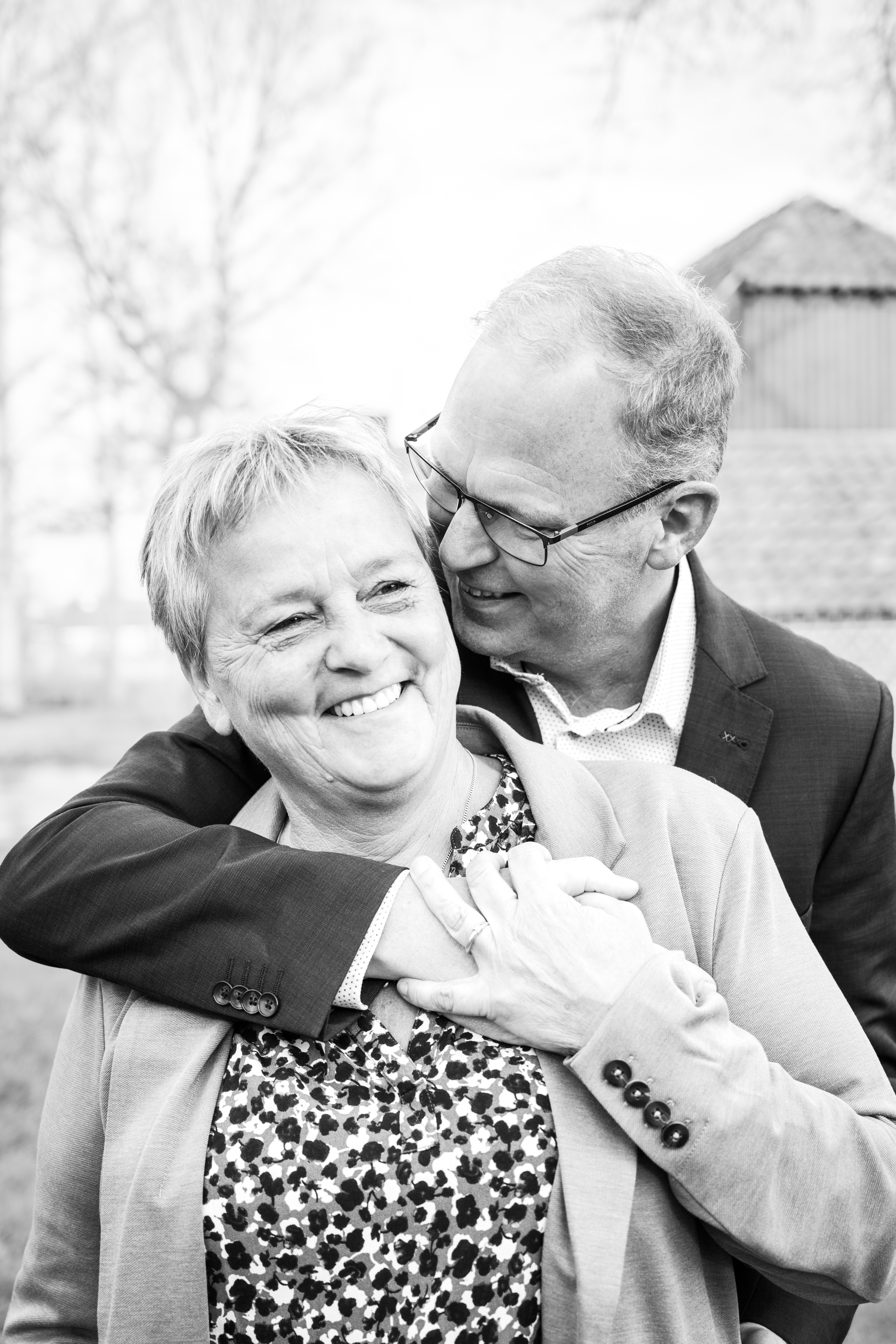 Jan stands behind Elke with his arm around her shoulder. She takes his hand in her hand and they are both smiling