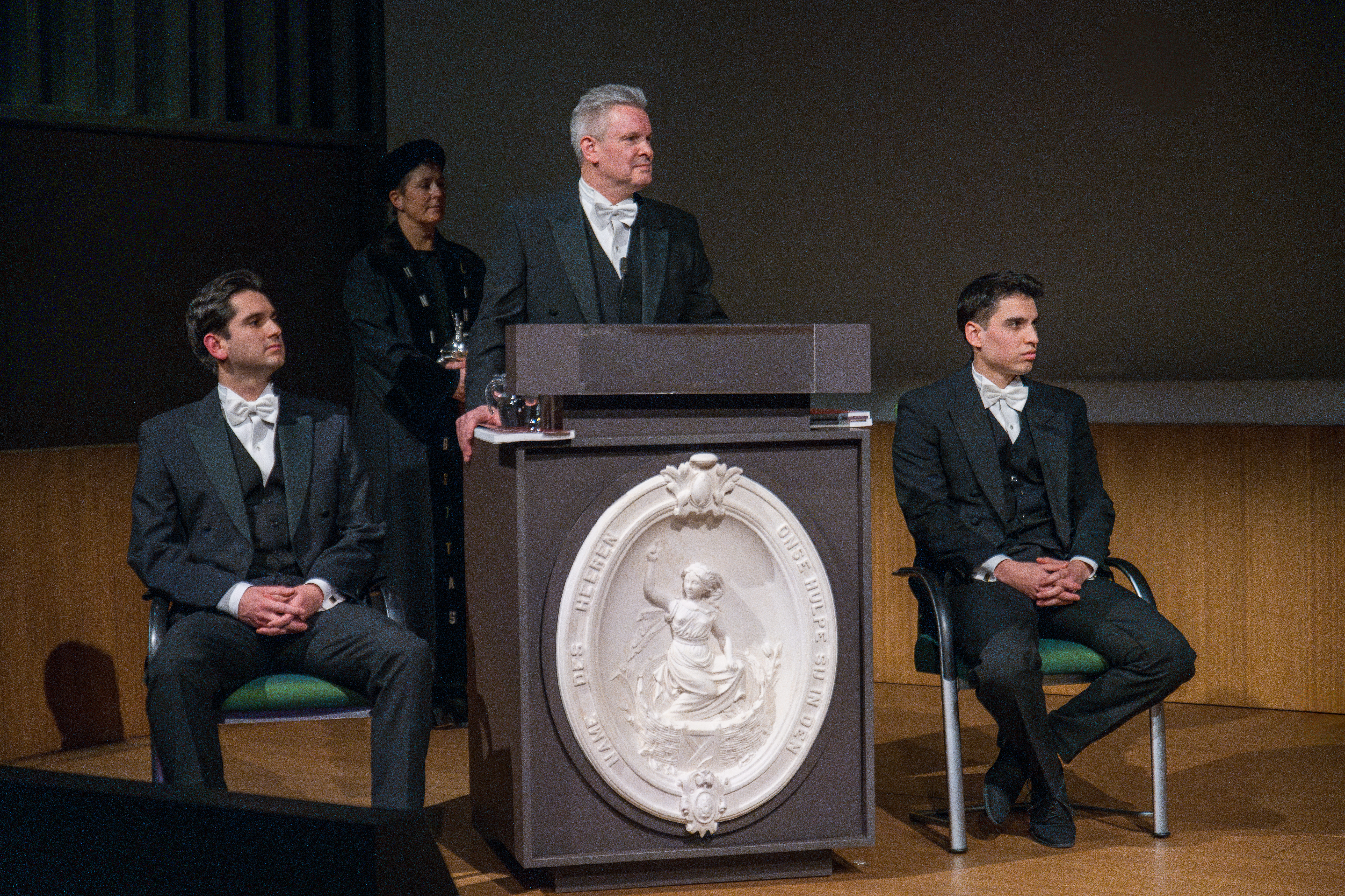 A man wearing a tuxedo with white bow tie stands behind a podium ready to defend his thesis and is flanked by two paranymphs